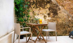 Un pequeño espacio al aire libre con dos sillas blancas y una mesa de madera, frente a una pared de piedra con vegetación.