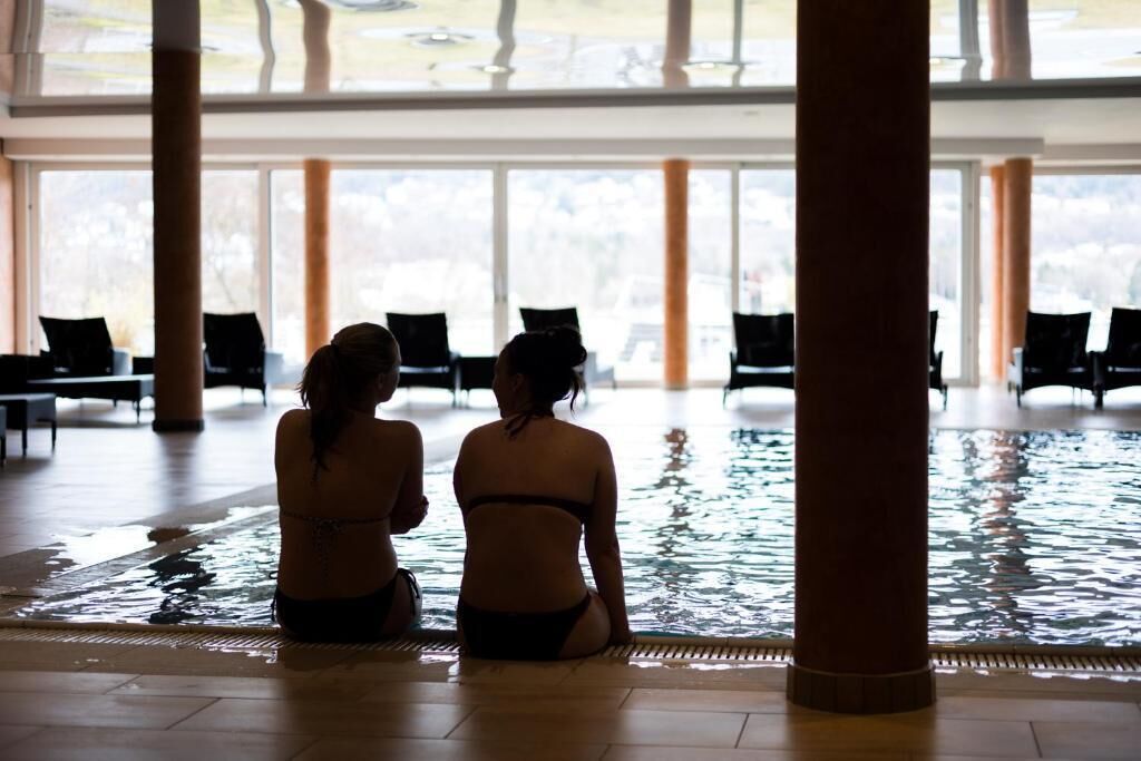 Two women sitting by a pool with chairs and large windows in the background.