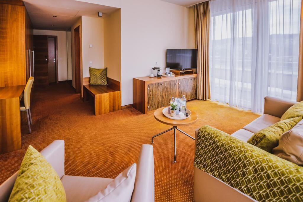 Une chambre d’hôtel avec un canapé, une petite table avec un plateau, un téléviseur posé sur un meuble en bois et une vue sur l’extérieur grâce à de grandes fenêtres.