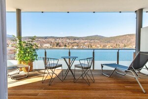 A balcony with a wooden floor, two chairs, a table, and a lounge chair, overlooking a lake and a town.