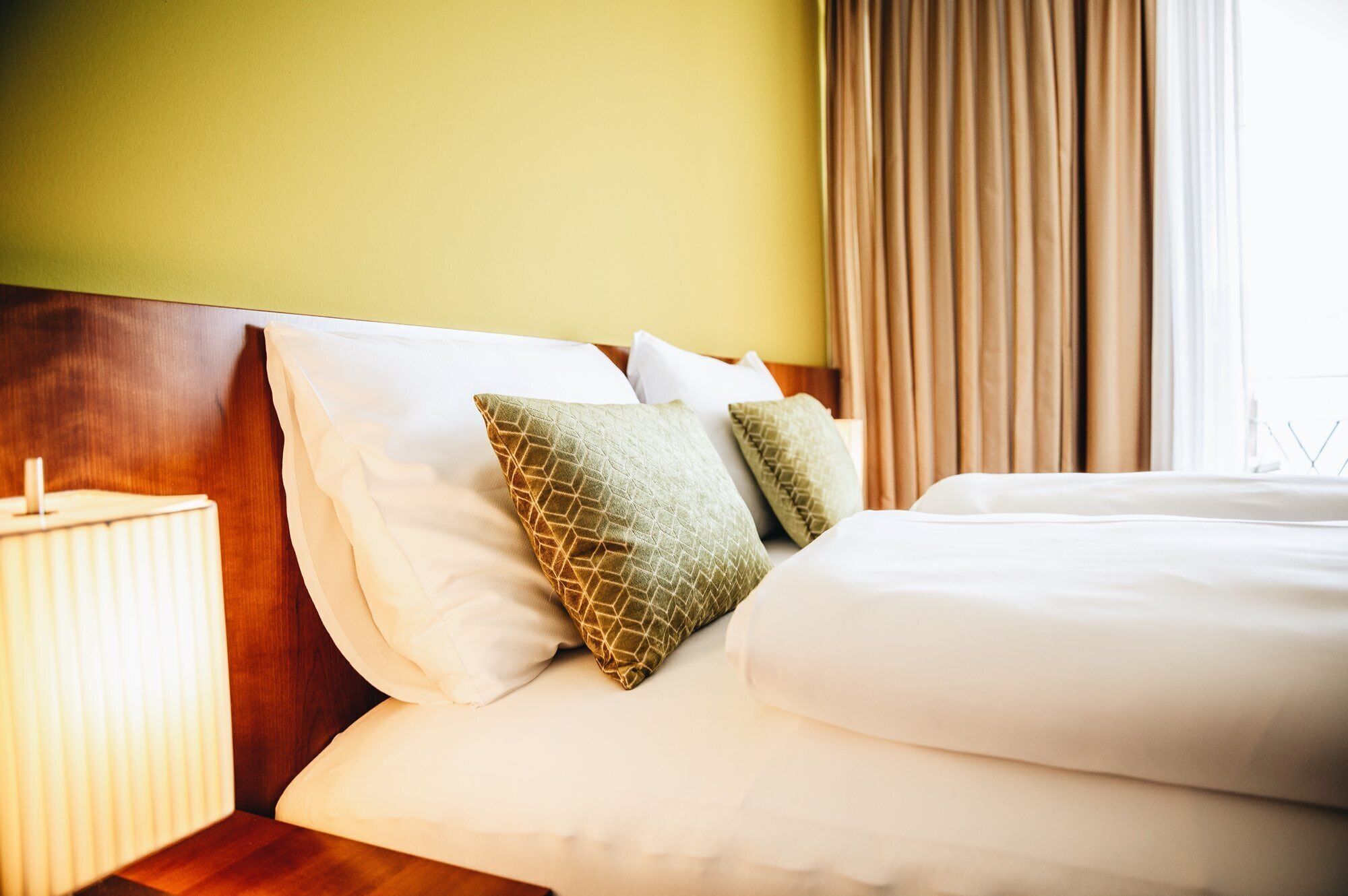 A neatly made bed with white linens and two patterned pillows, a wooden headboard, and a bedside lamp.