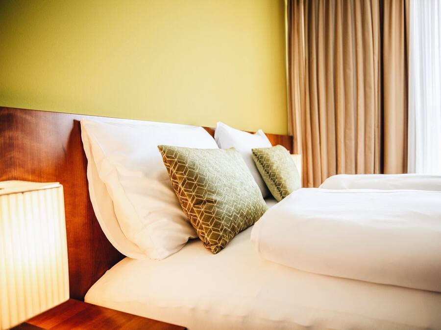 A neatly made bed with white linens and two patterned pillows, a wooden headboard, and a bedside lamp.