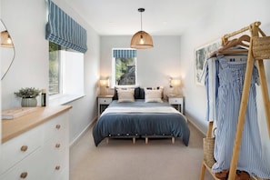 Une chambre à coucher comprenant un lit, des tables de chevet, un luminaire suspendu, une fenêtre avec des rideaux et un portant à vêtements.