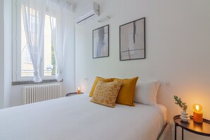 A neatly made bed with white and mustard pillows, a bedside table with a lamp and a small plant, and two abstract paintings on the wall.