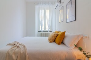 A neatly made bed with a white duvet and two pillows, one yellow and one white, in a room with white walls and a window with sheer curtains.