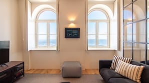A living room with two large arched windows overlooking the sea, a small ottoman, a black sofa with striped pillows, and a mounted TV.