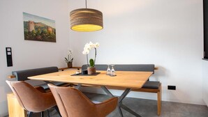 Un comedor con una mesa de madera, cuatro sillas marrones, una lámpara colgante de fibras naturales, un banco y una orquídea en maceta.