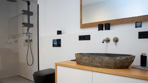 Un baño moderno con un lavamanos grande de piedra, un tocador de madera, un inodoro negro, una ducha con cabezal de ducha de mano y un espejo sobre el lavamanos.