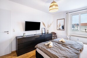 Une chambre moderne avec un lit, une commode, un téléviseur et une fenêtre donnant sur des immeubles.
