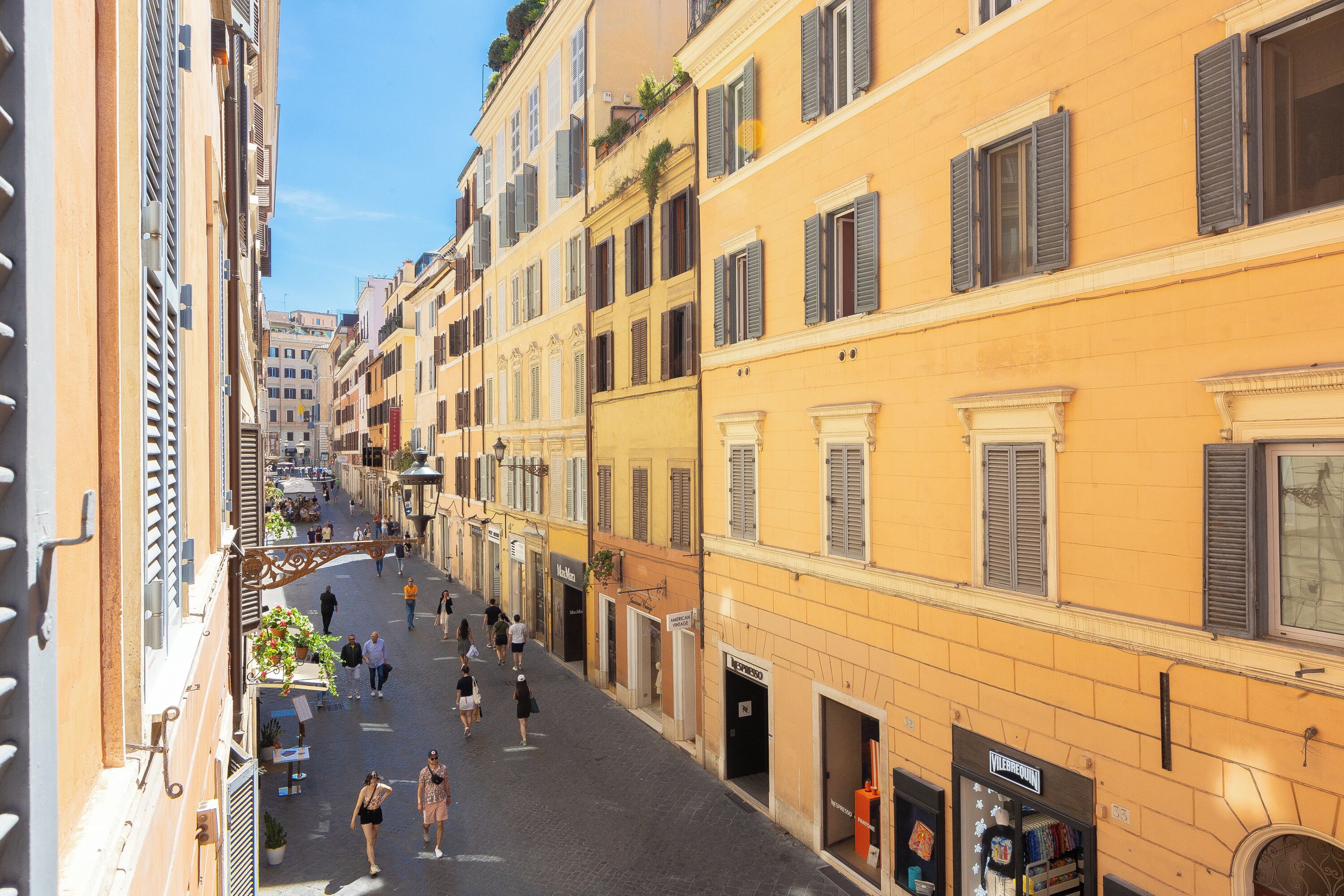 A narrow street lined with yellow buildings, some with shutters, and a few balconies with plants.