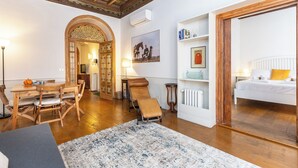 A room with a wooden dining table, chairs, a sofa, a patterned rug, a bed with white bedding, a radiator, and a bookshelf with books and decorative items. - Rental in Rome Frattina Atmosphere Apartment (Rome)
