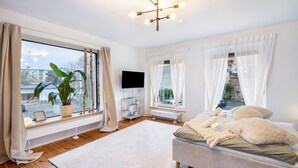 Un dormitorio moderno con un ventanal grande, un televisor de pantalla plana, una cama con almohadas mullidas y una alfombra blanca.