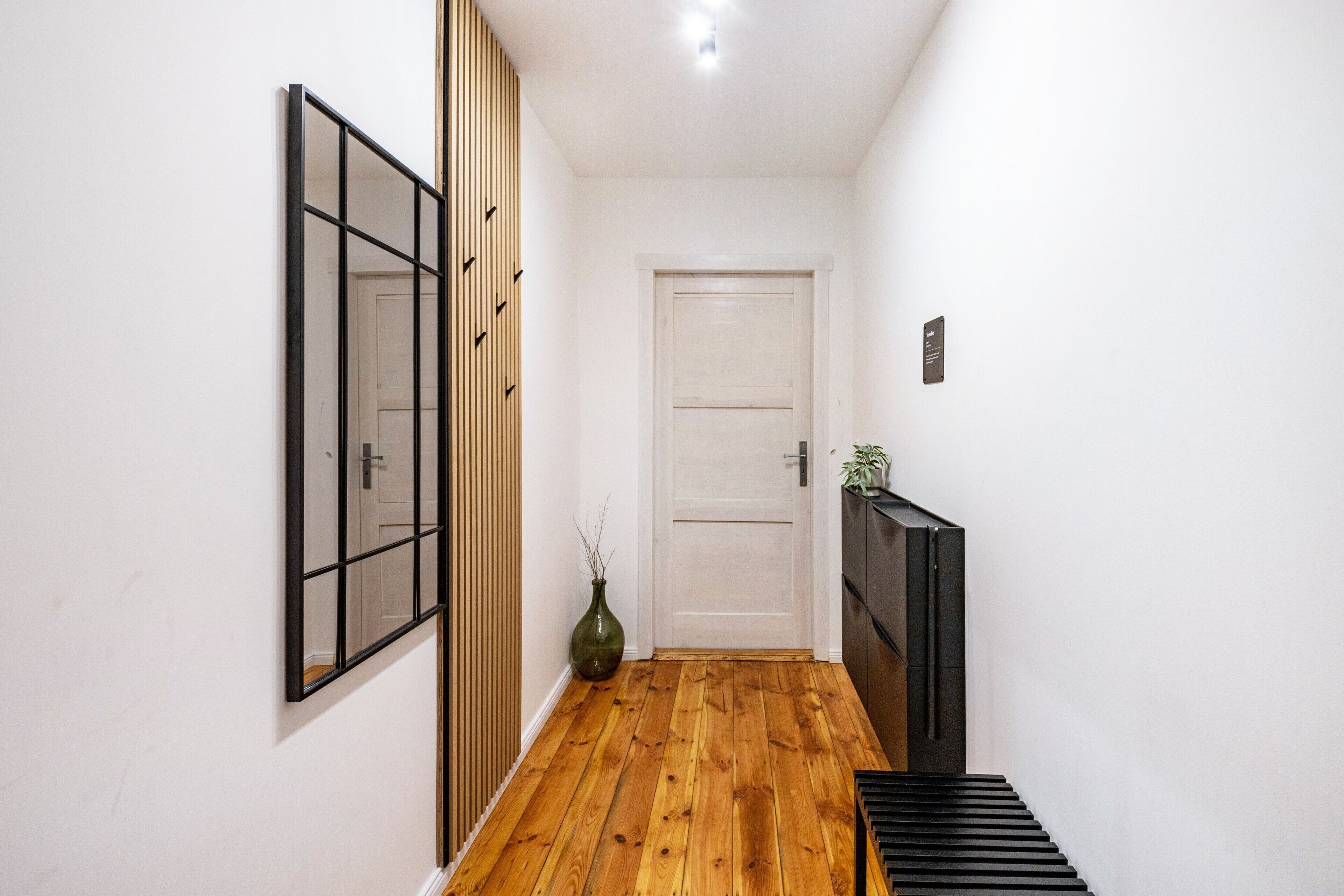 Un couloir avec un plancher en bois, une porte blanche, un miroir à cadre noir et une petite plante.