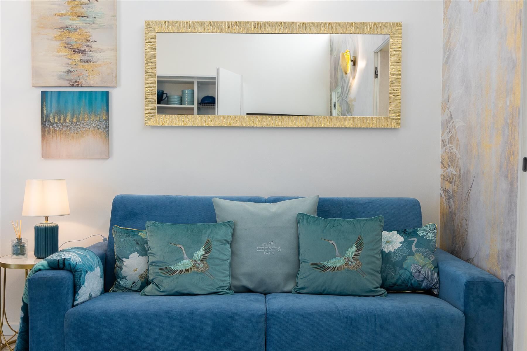 A blue sofa with three cushions, a gold-framed mirror, and a painting on the wall.