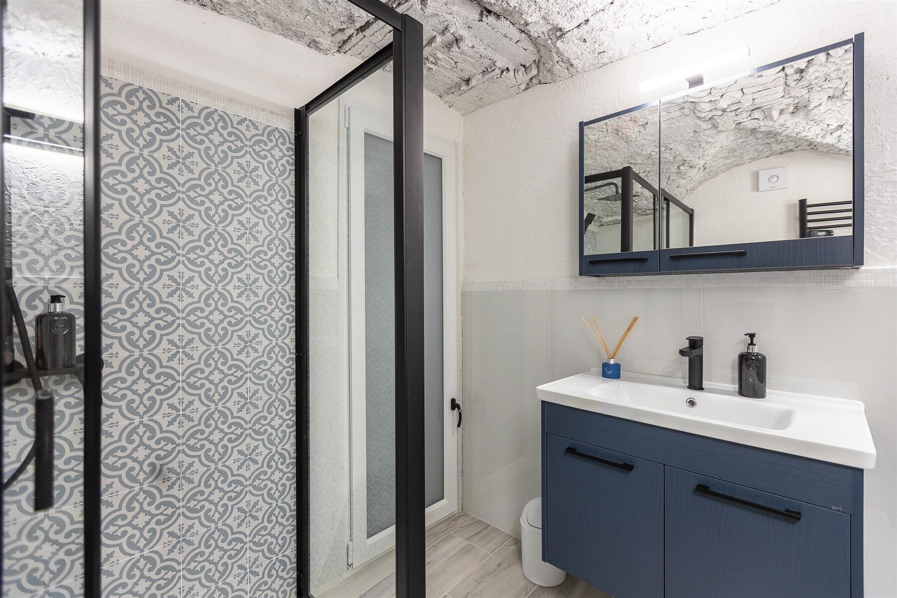Une salle de bain avec des carreaux décorés, un lavabo blanc, des meubles bleu foncé et un miroir qui reflète un escalier.