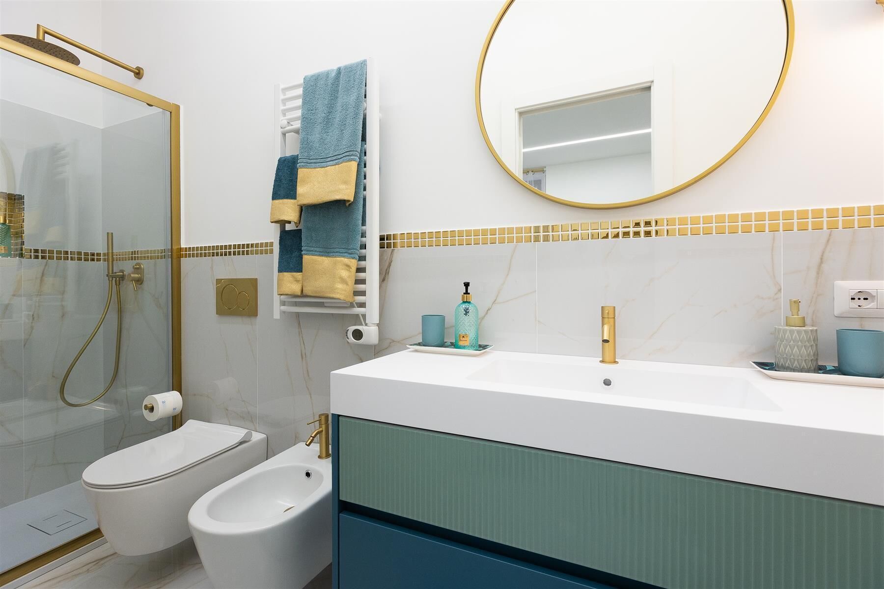 A modern bathroom with a glass-enclosed shower, a white bidet, a gold showerhead, a white sink with a gold faucet, and a mirror with a gold frame.