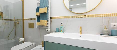 A modern bathroom with a glass-enclosed shower, a white bidet, a gold showerhead, a white sink with a gold faucet, and a mirror with a gold frame.