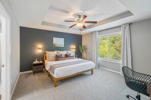 A bedroom with a bed, bedside tables, a ceiling fan, a window with curtains, and a view of greenery outside.