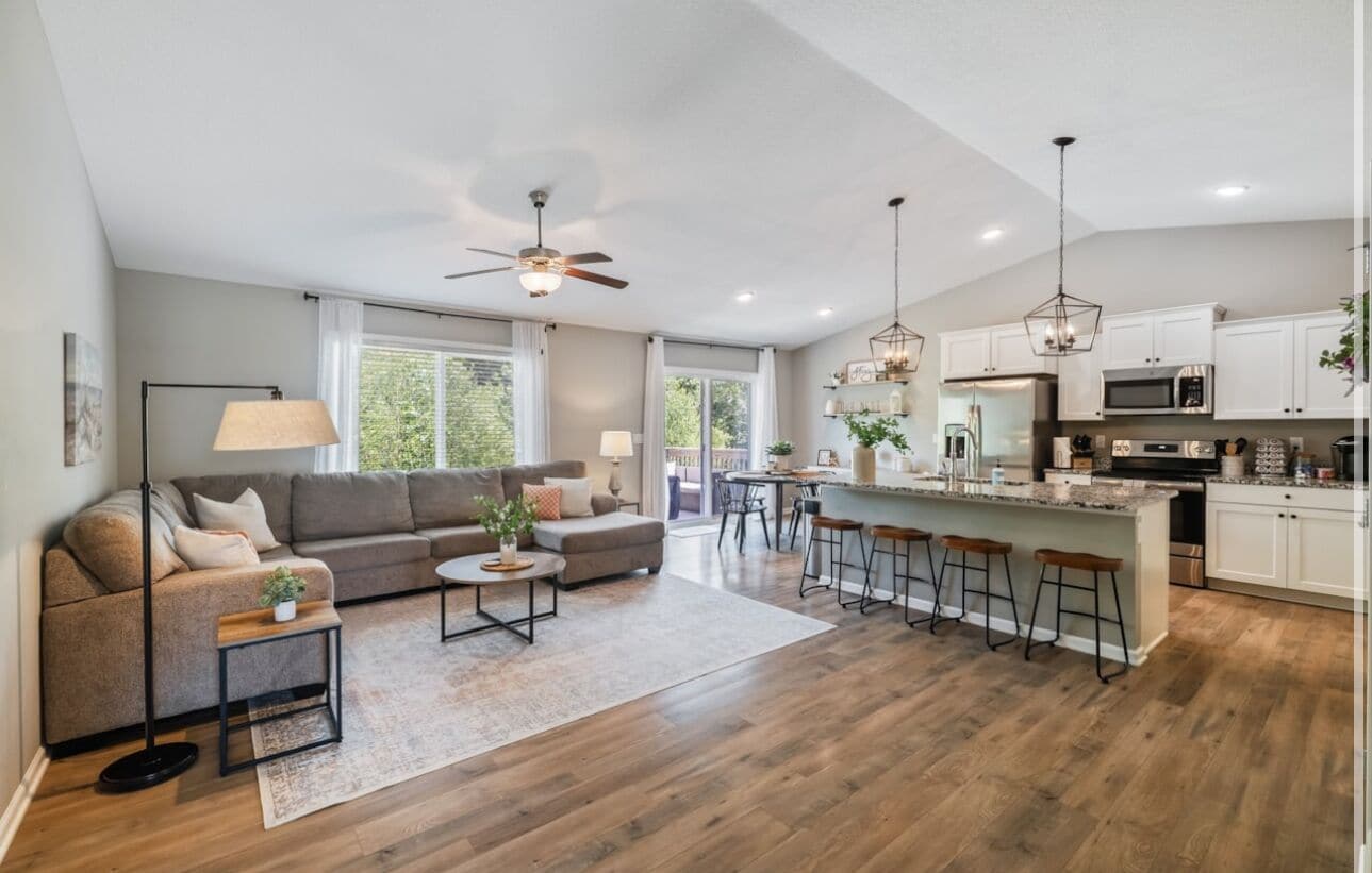 A spacious living room with a sofa, coffee table, and a dining area with bar stools. The kitchen features white cabinets and stainless steel appliances.