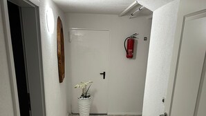 A hallway with a wooden floor, white walls, a white door, a white vase with flowers, and a fire extinguisher on the wall.
