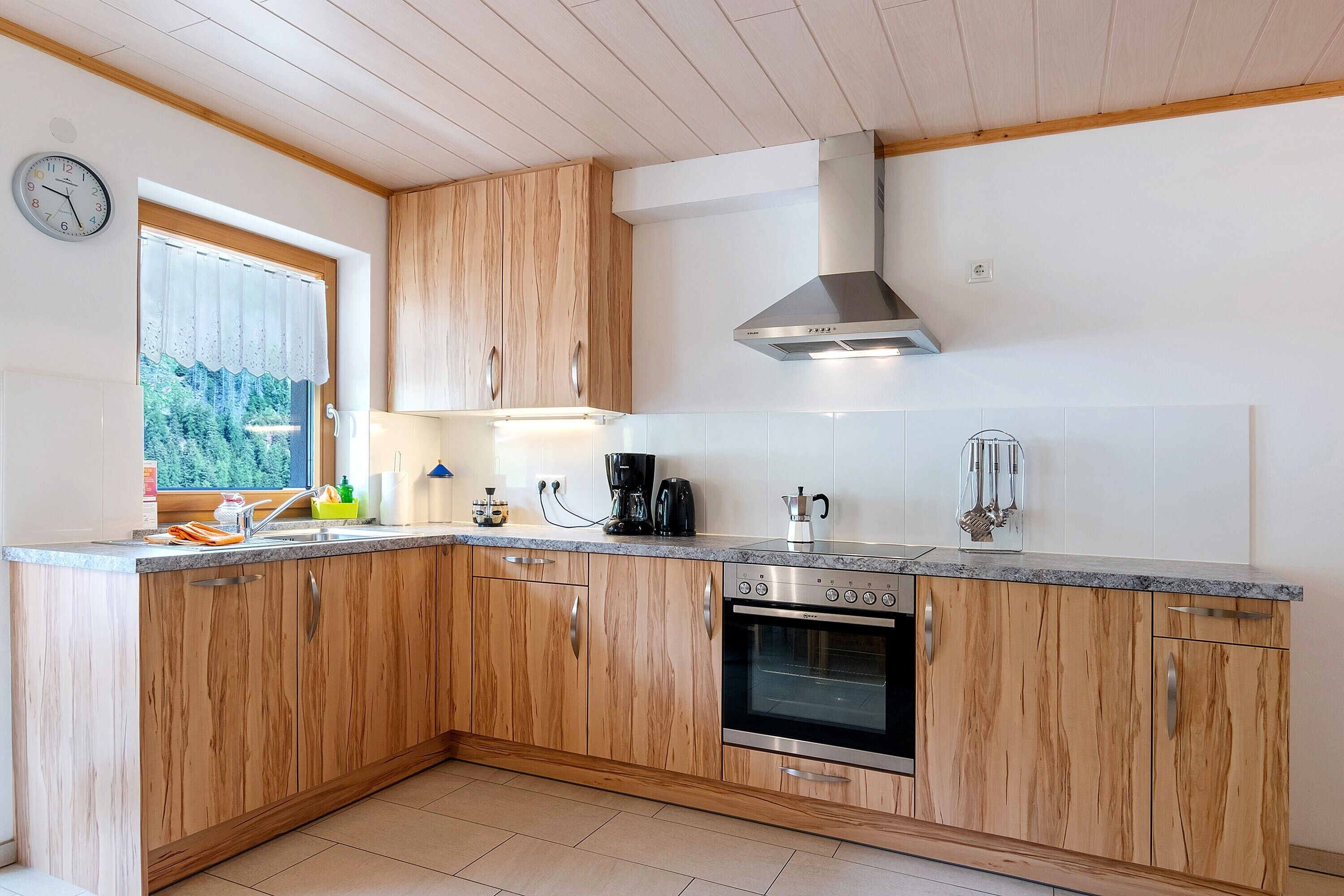 Una cocina con armarios de madera, horno empotrado, cafetera y ventana con vistas a la vegetación.