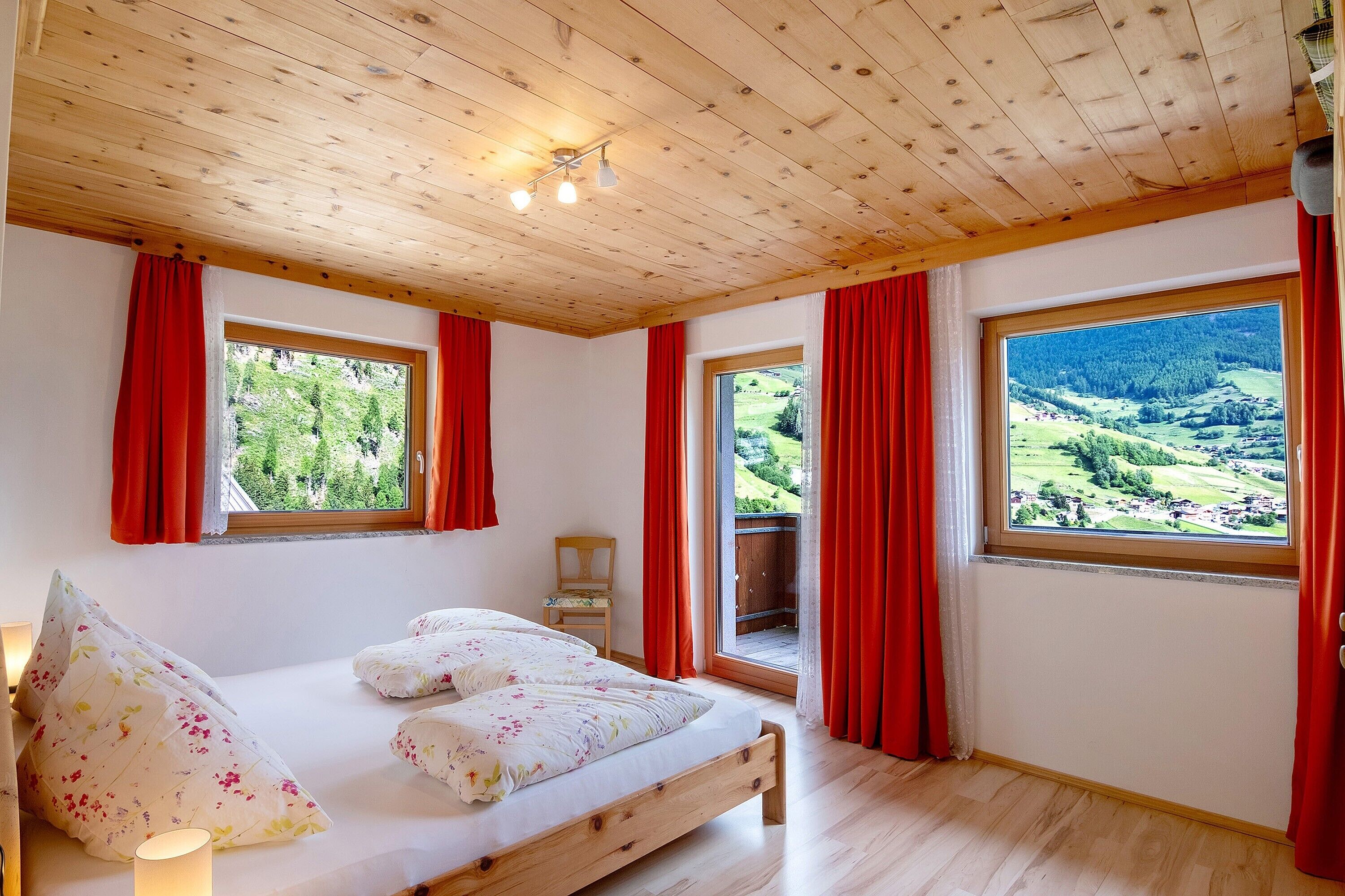 Un dormitorio con techo de madera, dos ventanas con cortinas rojas, una cama con ropa de cama floral y vistas a un paisaje verde.