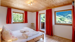 A bedroom with a wooden ceiling, two windows with red curtains, a bed with floral bedding, and a view of a green landscape.