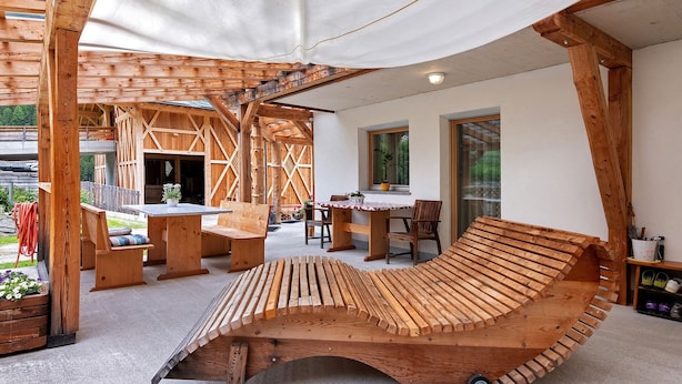 A wooden outdoor seating area with benches and a table, a wooden structure with a sloped roof, and a window with a view of greenery.