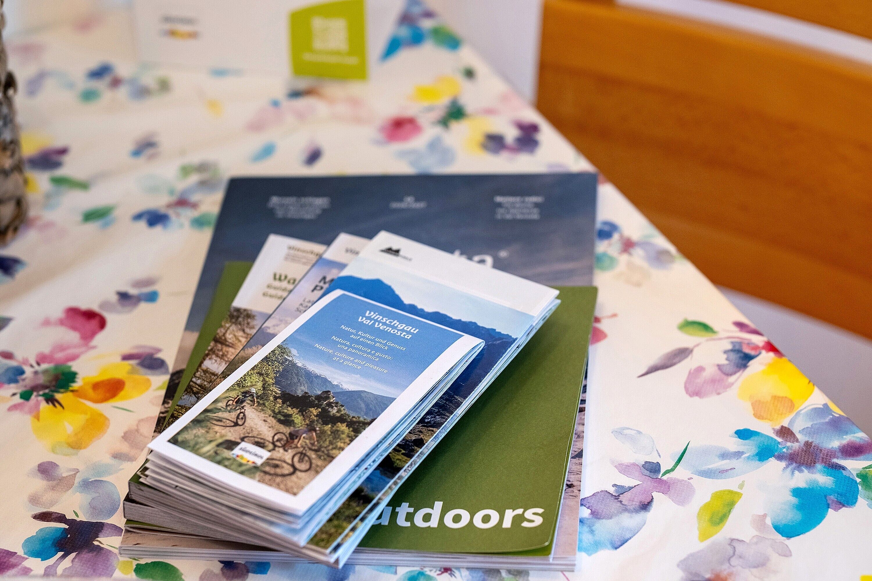 A stack of brochures with a mountain landscape cover, placed on a table with a floral tablecloth.