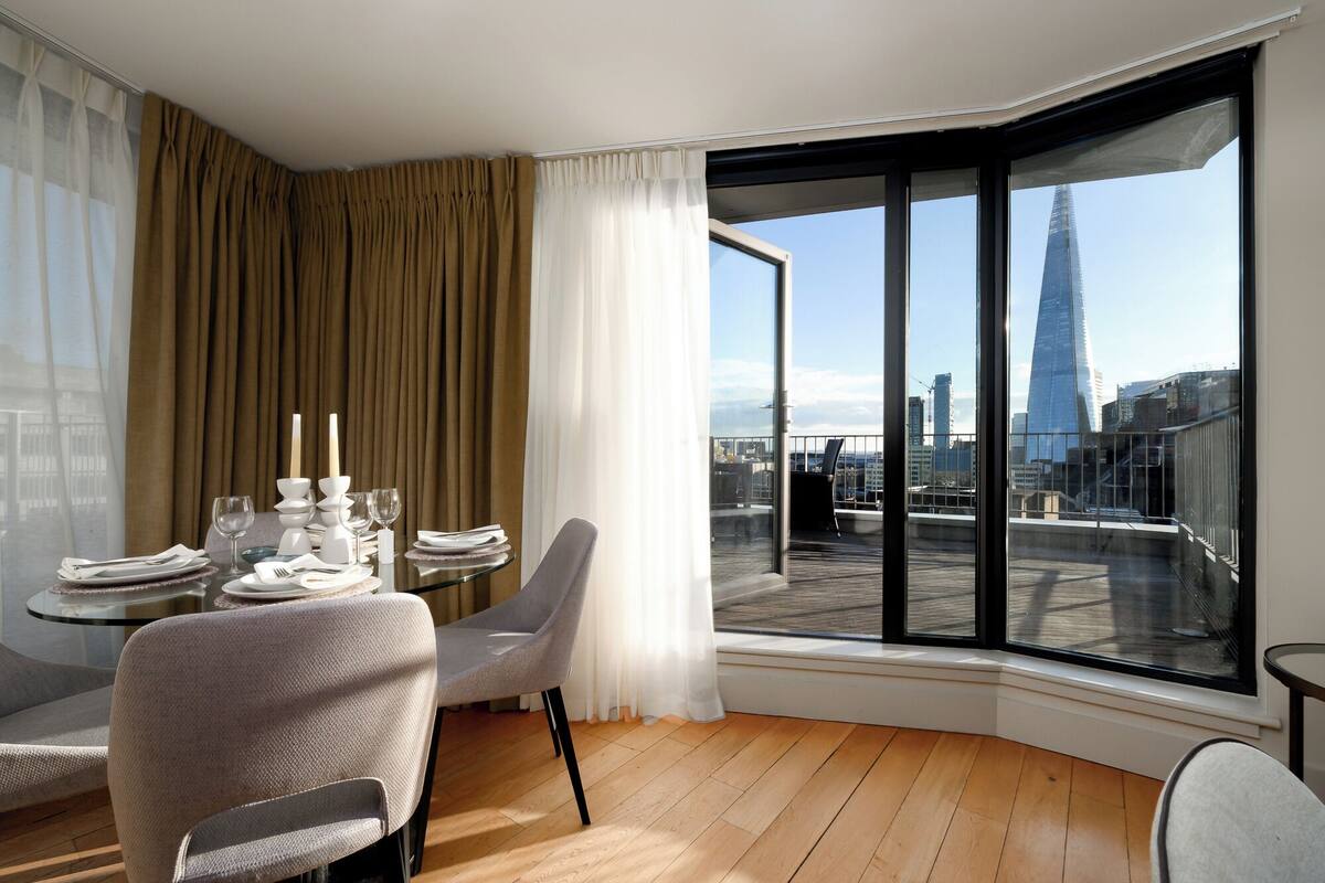 Een hoekappartement met een eettafel gedekt voor vier personen, een groot raam met uitzicht op een hoog gebouw, en een balkon met een stoel.