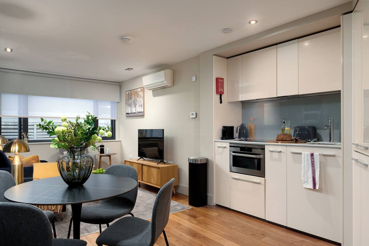Een moderne keuken met witte kasten, een ingebouwde oven en een magnetron. Er is een eethoek met een ronde tafel en stoelen, en een vaas met groene planten.