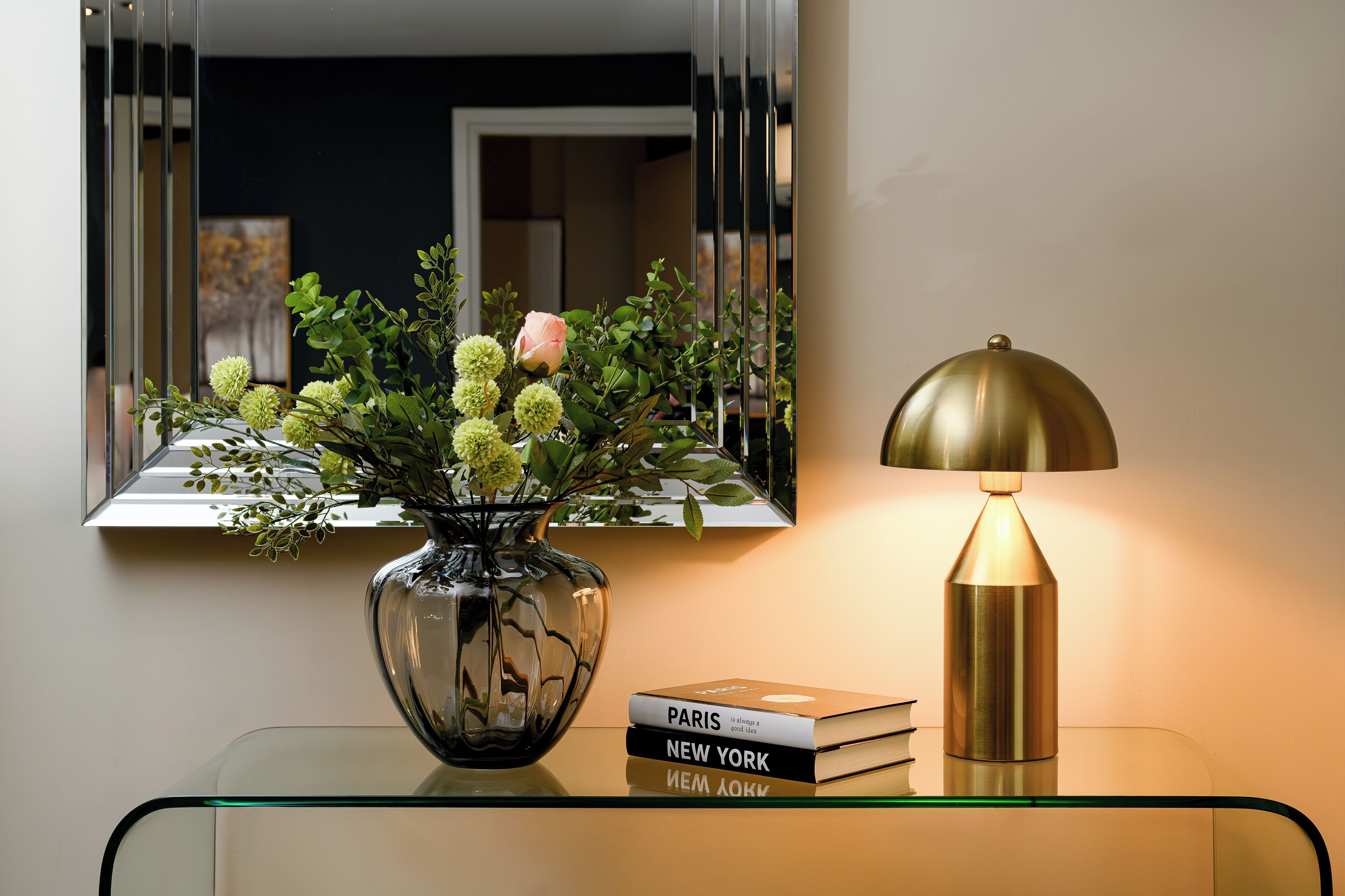 Ein Glastisch mit einer Blumenvase, einem Buch mit dem Titel „Paris, New York“ und einer Lampe.