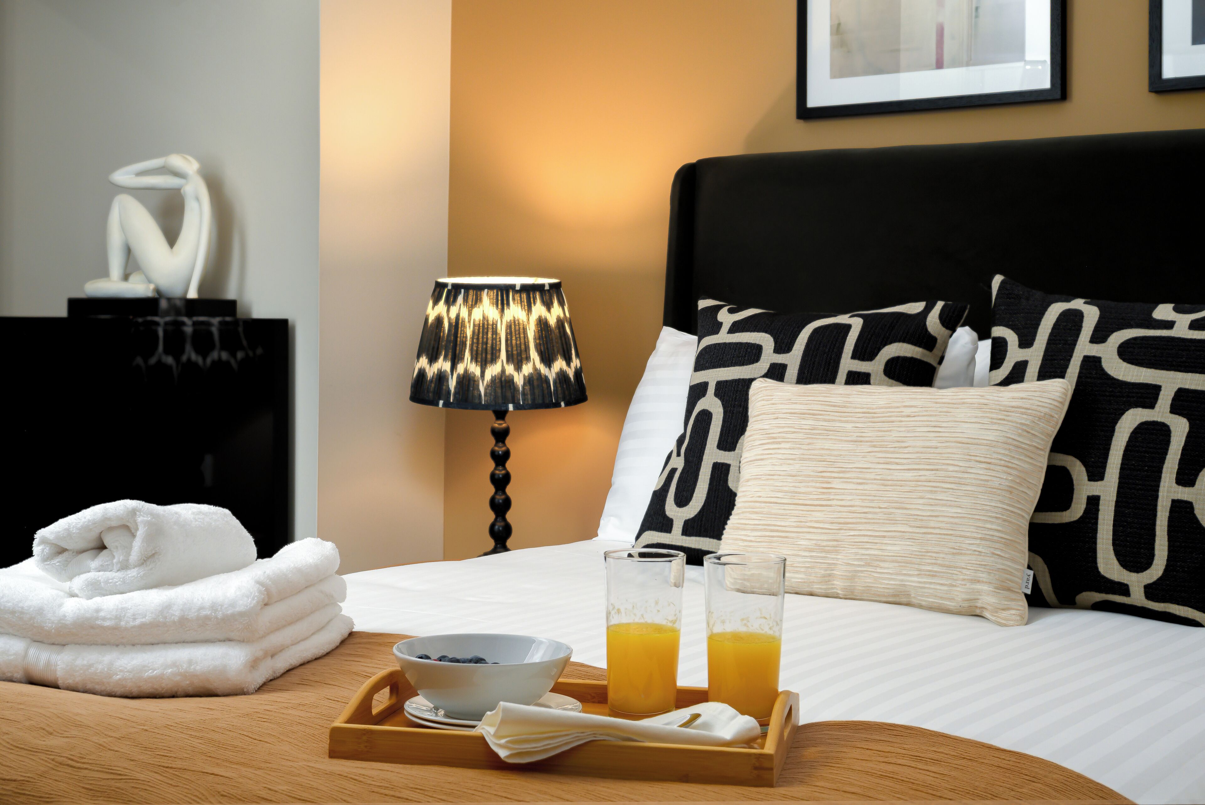 A neatly made bed with black and white patterned pillows, a bedside lamp, and a tray with a bowl of fruit and two glasses of orange juice.