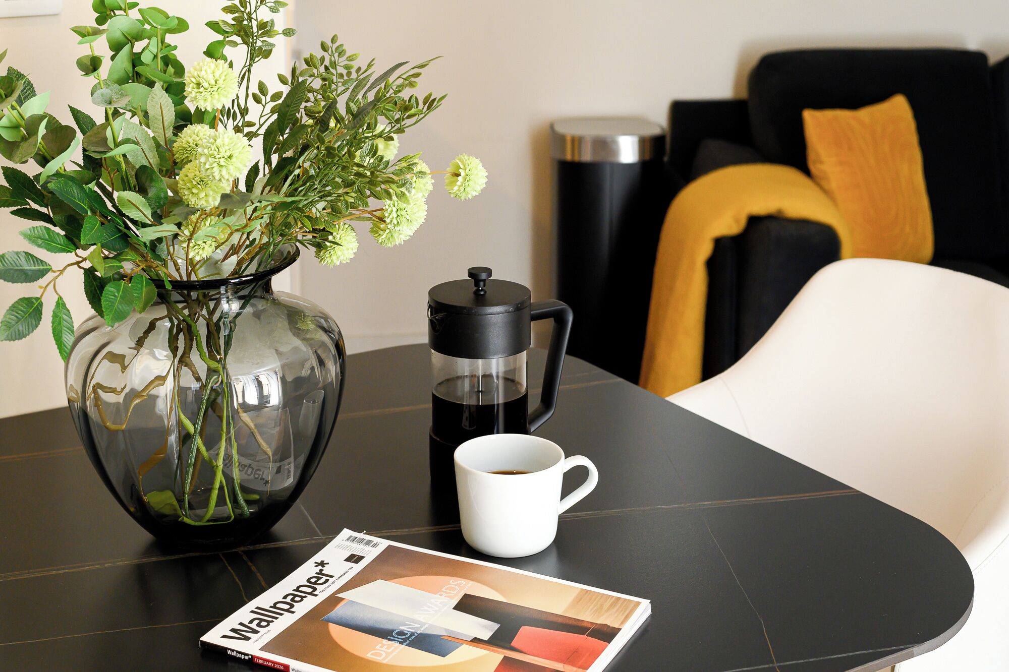 Una mesa con una revista, una cafetera y un florero con flores.