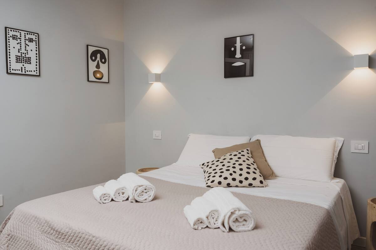 A neatly made bed with white linens and rolled towels, a polka-dotted pillow, and two wall-mounted light fixtures.