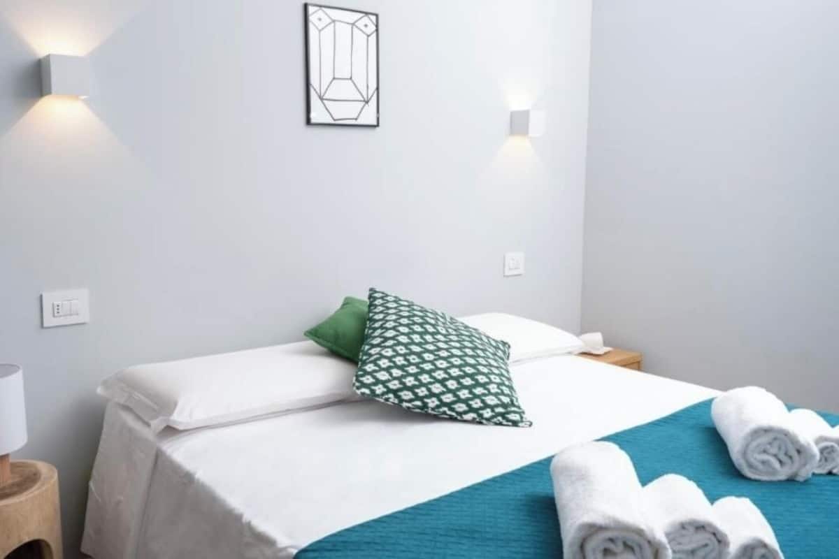 A hotel room with a bed, a bedside table with a lamp, a wall-mounted light, and a framed picture.
