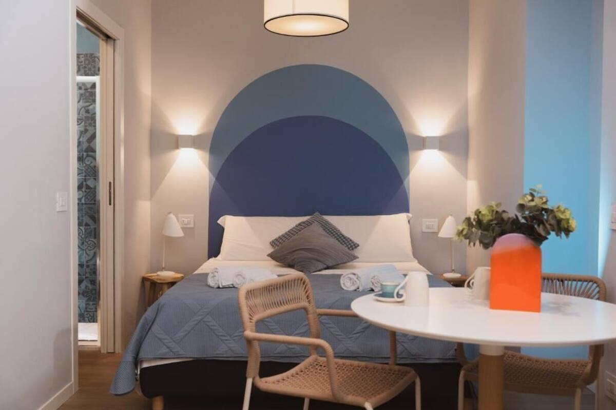 A bedroom with a bed, two bedside lamps, a round table with a vase, and a chair.