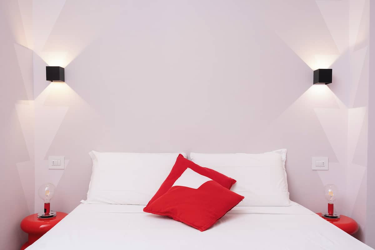 A bed with white bedding, a red pillow, and two bedside tables with red bases and clear glass lamps.