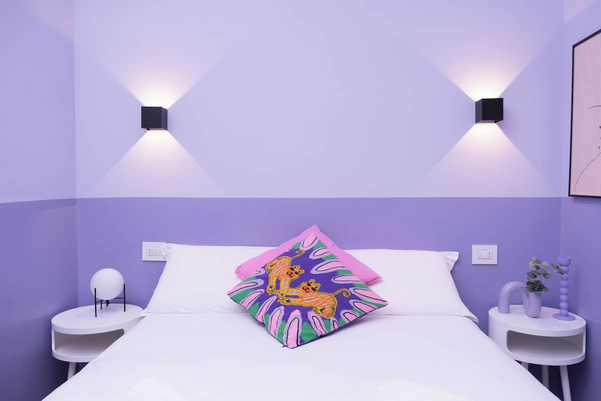 A bedroom with a bed, two nightstands, a wall-mounted light fixture, and a decorative pillow.
