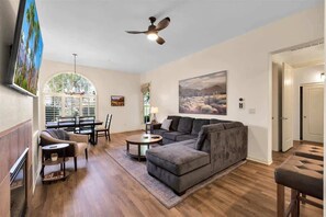 Ein geräumiges Wohnzimmer mit einer Eckcouch, einem Essbereich mit Tisch und Stühlen, einem Kamin und einem großen Fenster mit Jalousien.