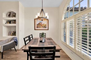 A dining area with a wooden table, chairs, a chandelier, and a window with blinds.