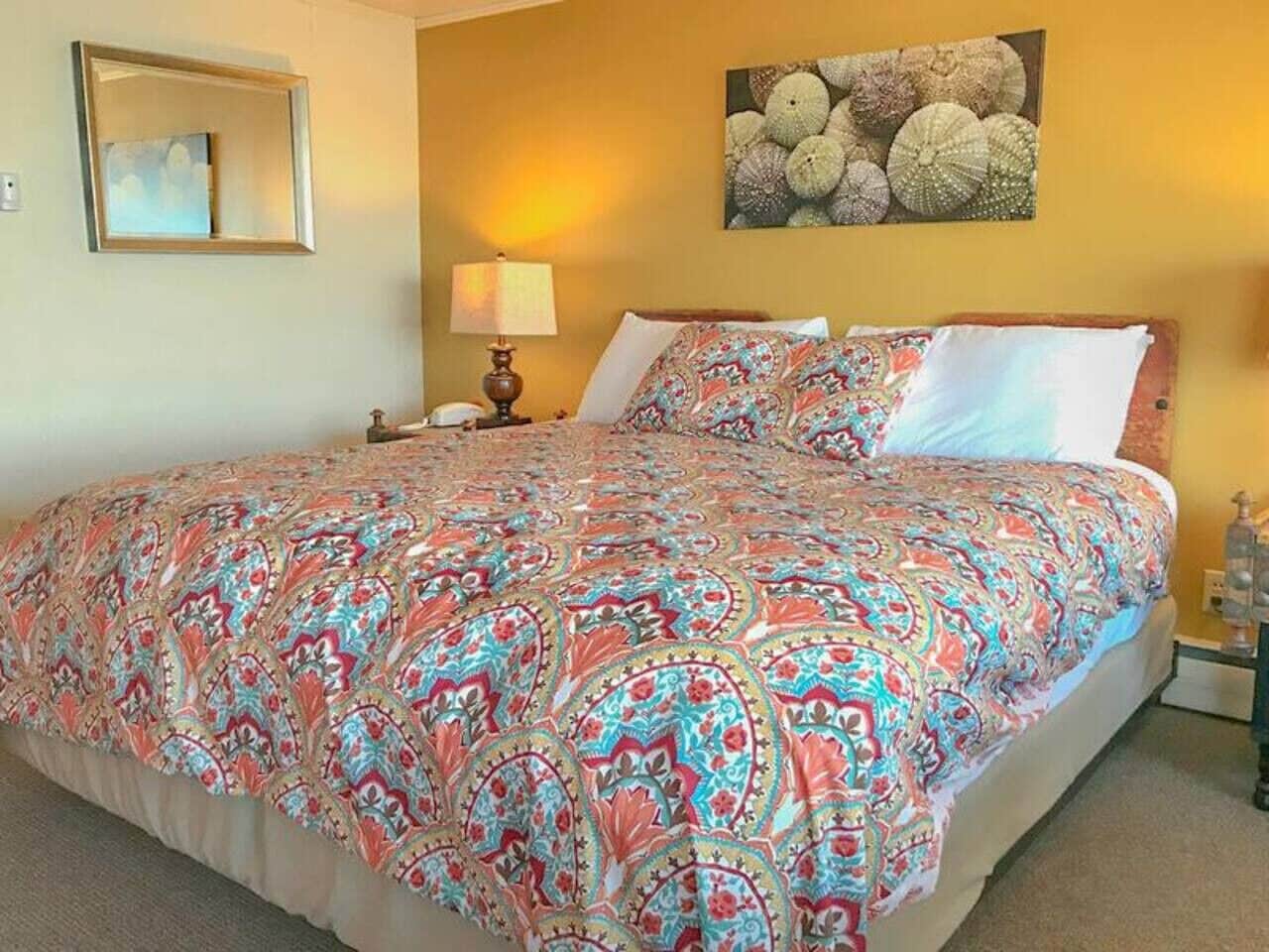 A large bed with a colorful patterned bedspread, a bedside table with a lamp, and a wall-mounted picture of seashells.