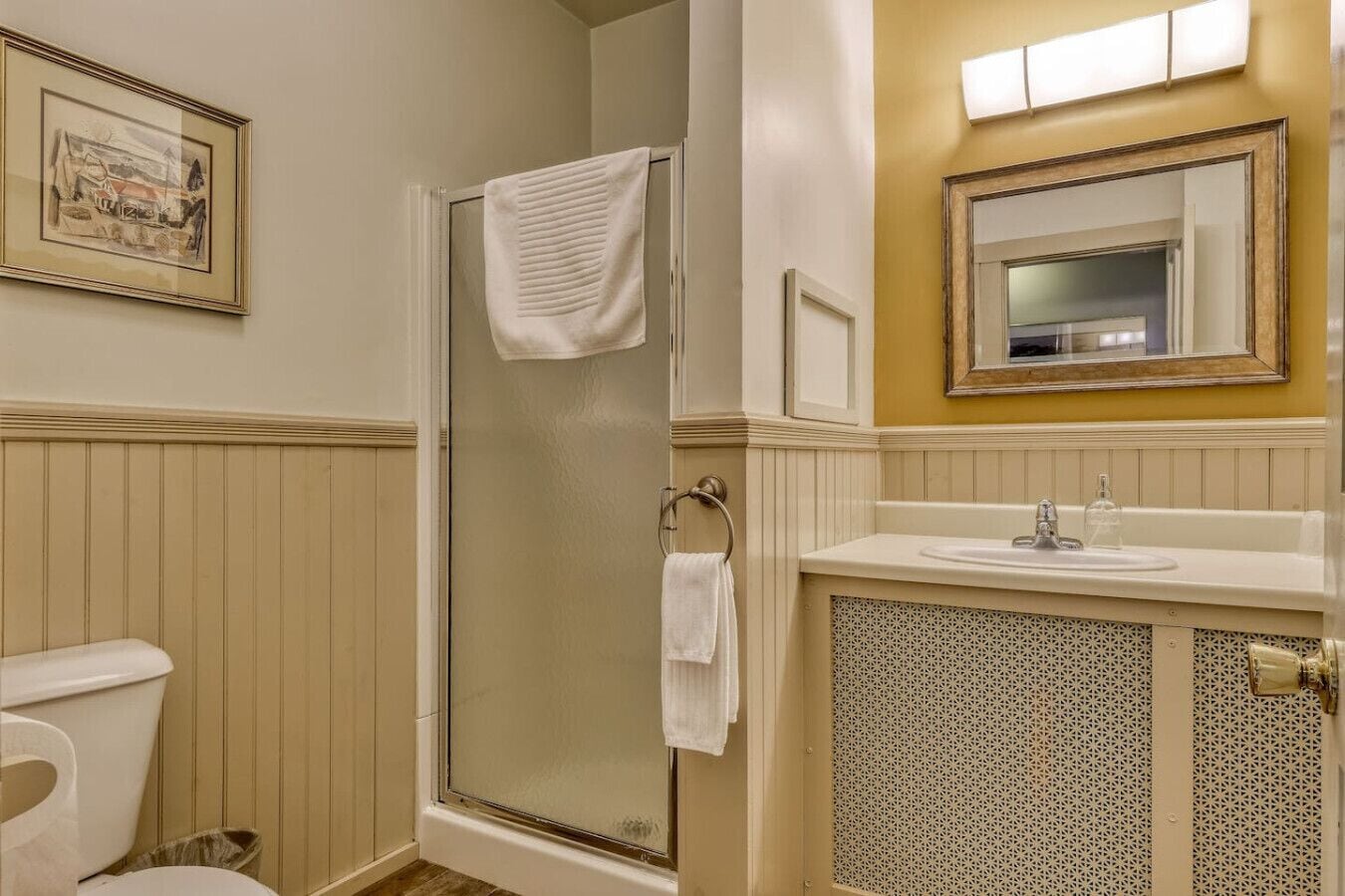 A bathroom with a glass shower enclosure, a white toilet, a sink with a mirror, and a framed picture on the wall.