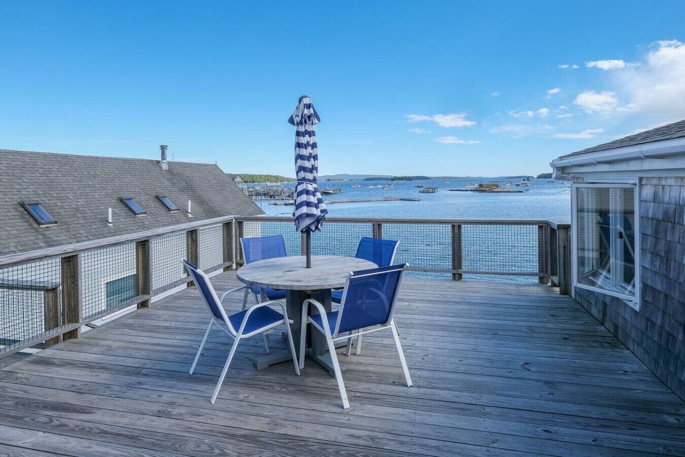 A wooden deck with a round table and blue chairs, overlooking a body of water with boats.