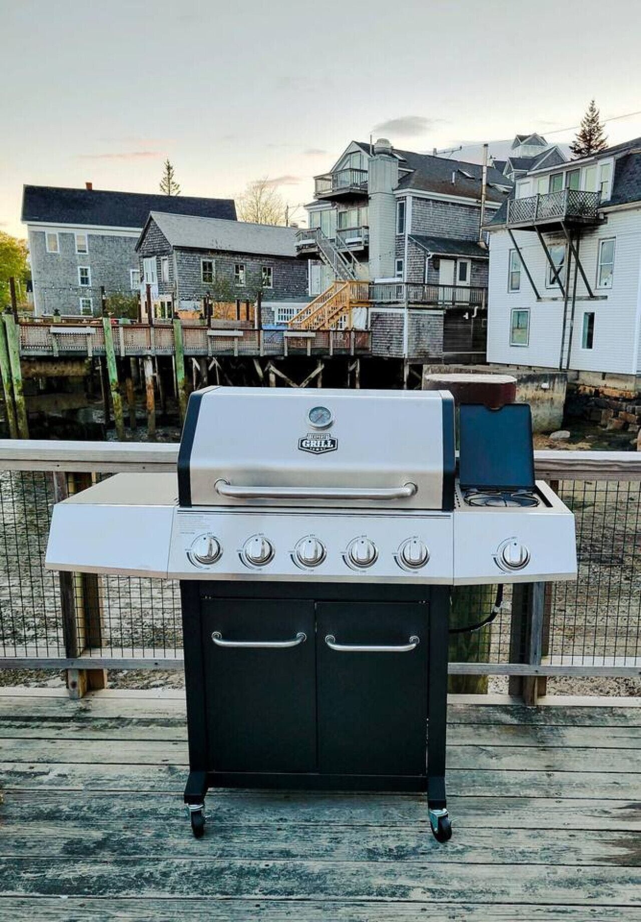A portable grill with five burners, set on a wooden deck with a view of residential buildings in the background.