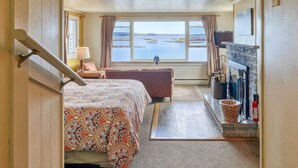 A bedroom with a bed, a small table with a lamp, a view of the sea, and a fireplace. - Inn on the Harbor (Stonington)
