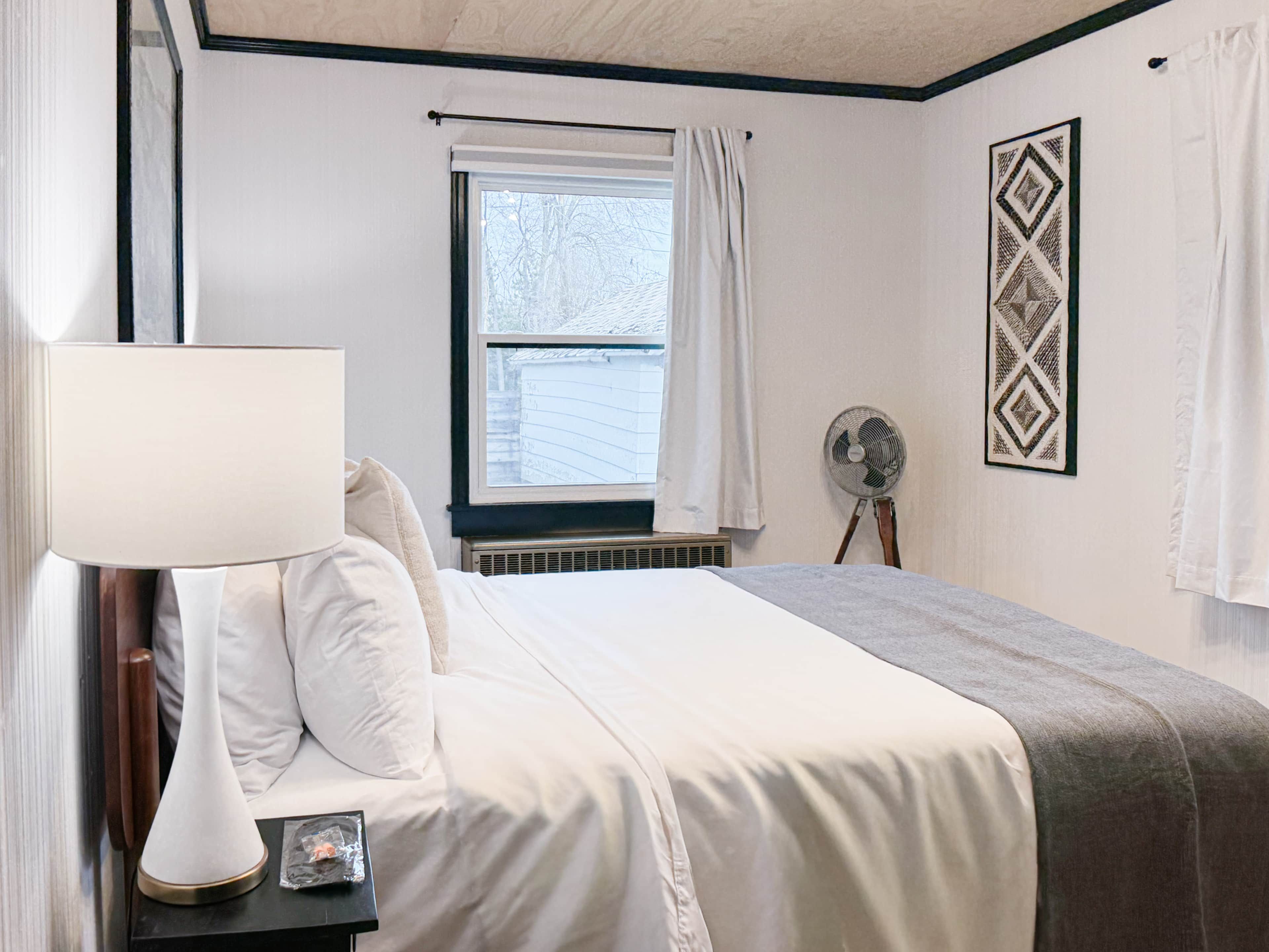 A bedroom with a bed, a bedside table with a lamp, a window with curtains, a wall-mounted fan, and a framed picture.