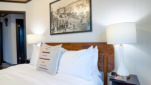 A hotel room with a bed, two bedside lamps, a nightstand, a framed historical photo on the wall, and a wooden headboard. - Stylish 1Br Near Main St | The Troubadour Suite (Sheridan)