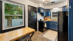 A compact kitchen with a dining table, black cabinets, a black refrigerator, and a window with a view of trees. - Stylish 1Br Near Main St | The Troubadour Suite (Sheridan)