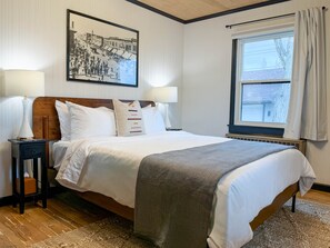 A neatly made bed with white linens, two bedside lamps, a framed black and white photo on the wall, and a window with curtains. - Stylish 1Br Near Main St | The Troubadour Suite (Sheridan)